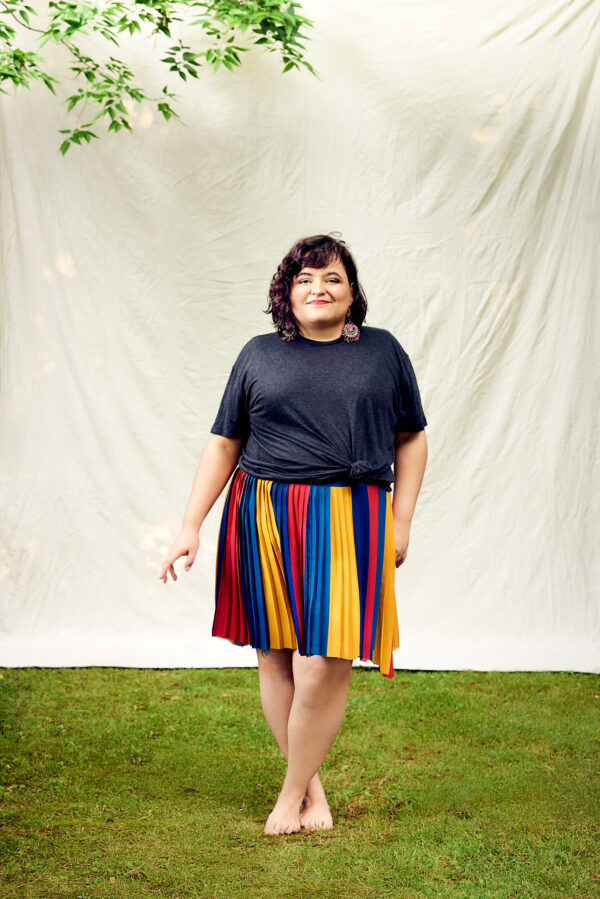 A full body shot of a woman, Aline Mello, standing barefoot on grass in front of a white drapery. She is wearing a blue short sleeve top and a blue, yellow, red pleated skirt.