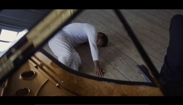 A man wearing white writhes on the floor in front of a piano.