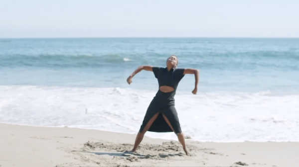 An individual standing on the sand at the beach, inches from the water, legs spread apart with arms out to the side and bent at the elbows. The person is looking up.