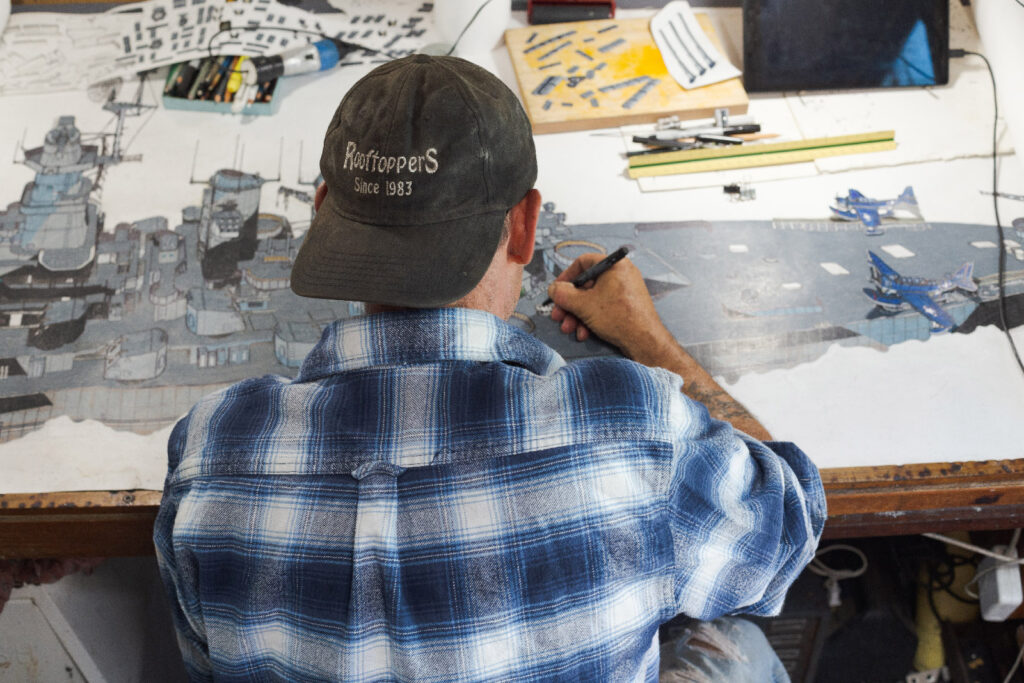Back view of artist JD Smith sitting and painting. He is wearing a blue and white flannel shirt with a baseball cap worn backwards.