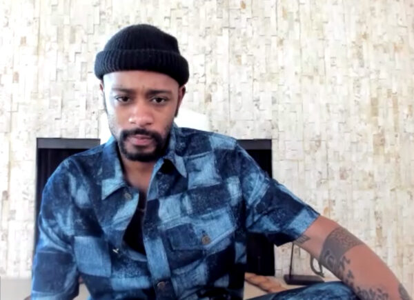 Image of Black American actor LaKeith Stanfield sitting on a chair in front of a white wall. He is wearing a blue skullcap and a checkered blue button down short sleeve shirt. He is looking directly into the camera.