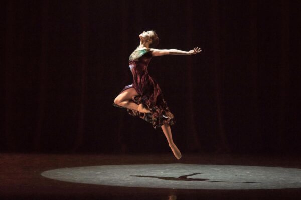 A dancer in a spotlight with her arms spread out and back, jumping in the air with one leg bent at the knee, her head tilted back.