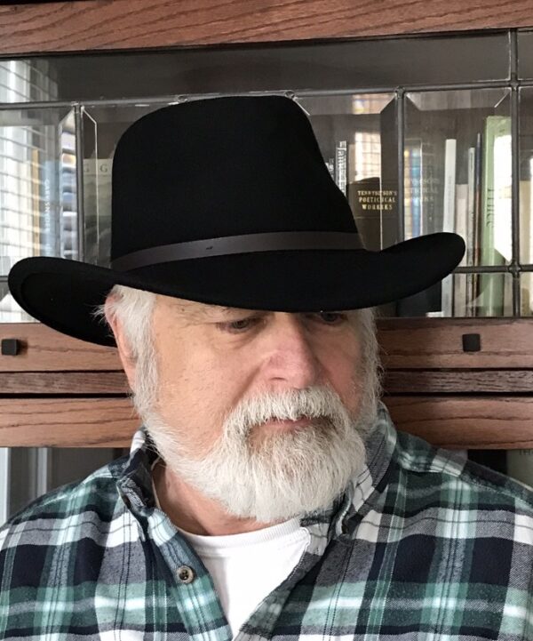 Image of the poet Michael Simms wearing a black cowboy hat and a blue and green flannel shirt. He is in front of a bookcase, looking off to his left.
