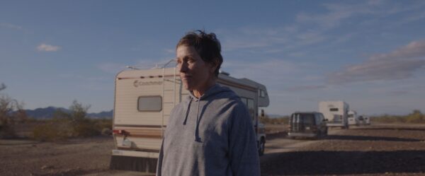A screenshot of actress Frances McDormand standing in front of a RV, parked on the side of the road under a blue sky, from the film Nomadland.