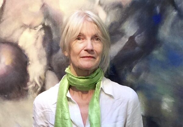 Image of poet Rachel Hadas looking into the camera, wearing a white top and a lime green scarf. She is standing in front of a painting.