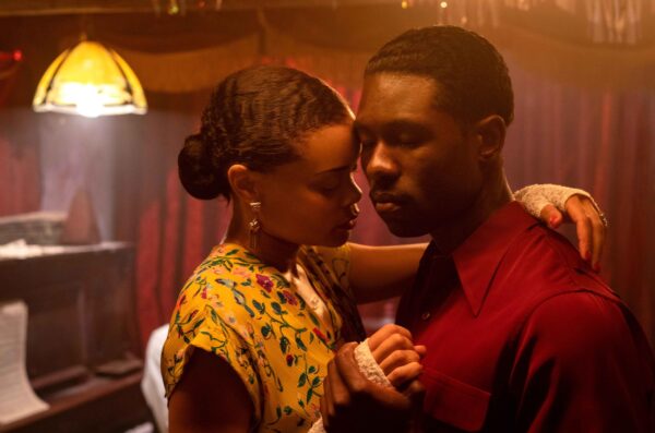 Image of a Black couple dancing. The Black American actress Andra Day is wearing a yellow flower print dress, her hair tied in a bun. The Black American actor Trevante Rhodes is in a red shirt. Both of their eyes are closed.
