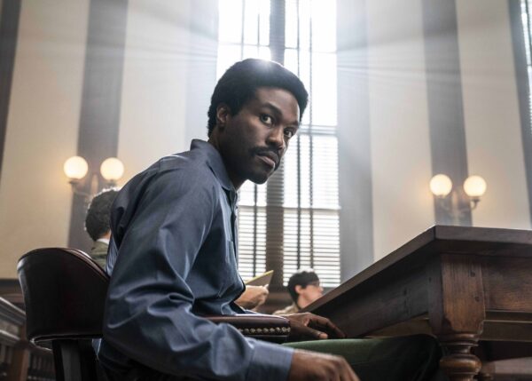 A Black man in a blue shirt turning to the camera, sits at a wooden desk, light coming in from the large window behind him.