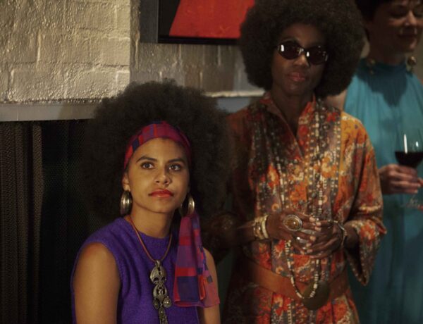 Image of Black American actress Zazee Beetz in a purple tank top and red head scarf. She is seated next to two men, all of them looking off to the left of the frame.