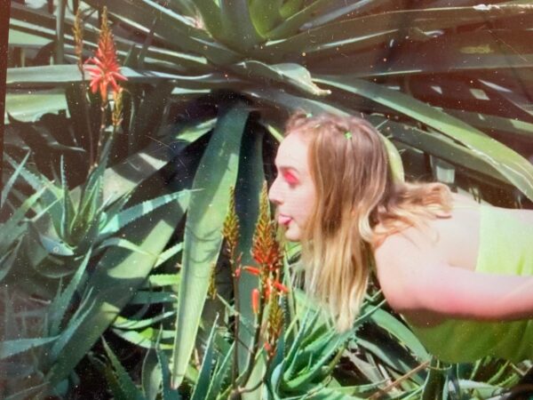 A woman leans toward a plant, sticking out her tongue as if she is an insect or reptile about to lick the leaf.