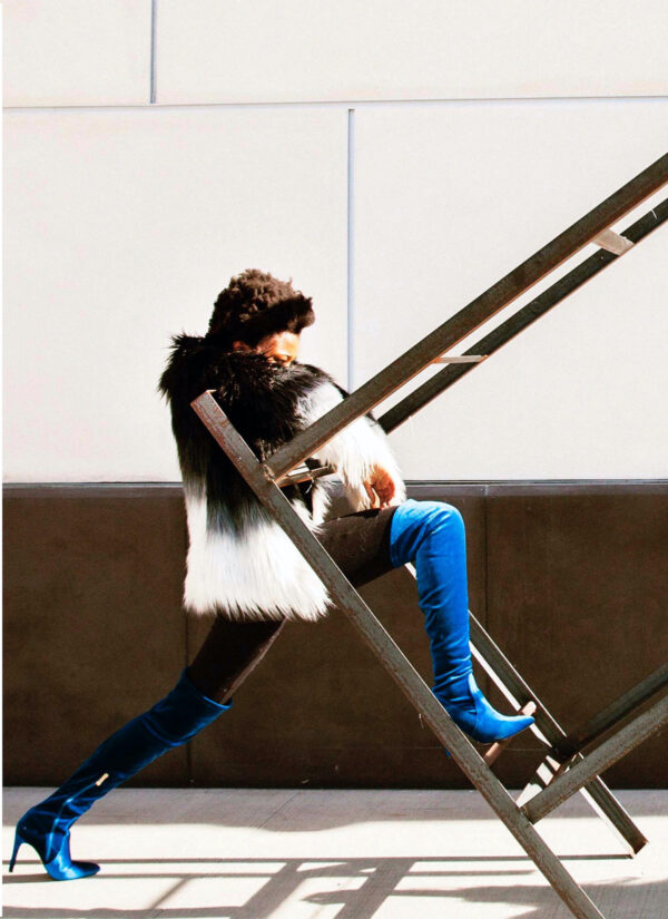 A dancer, Brianna Mims, straddles a tilted table. She wears bright blue high-heeled boots, tight blue jeans and a striking brown and white vest. Her face can't be seen, only the top of her head.