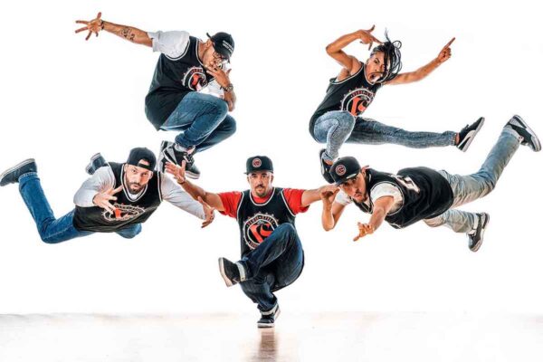 One dancer squats on one leg while four other dancers are in the air striking street dance poses.