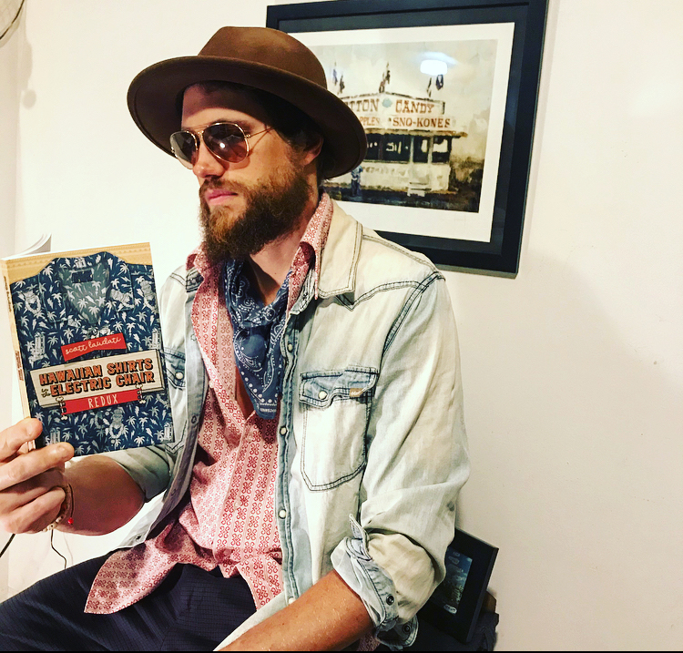 Scott Laudati, looking very cool in his dark shades, brown hat, blue scarf, and pink shirt is reading from his own book, Hawaiian Shirt in the Electric Chair REDUX.