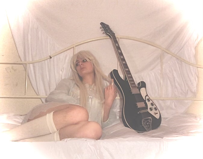 A white femme in a short white dress, a long blond wig, and a black electric guitar sitting on a bed.
