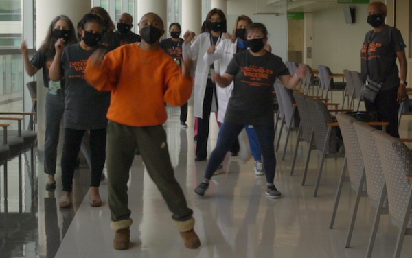 A group of people, some in hospital staff jackets dance in a room. All are masked and the film encourages vaccination against the virus.