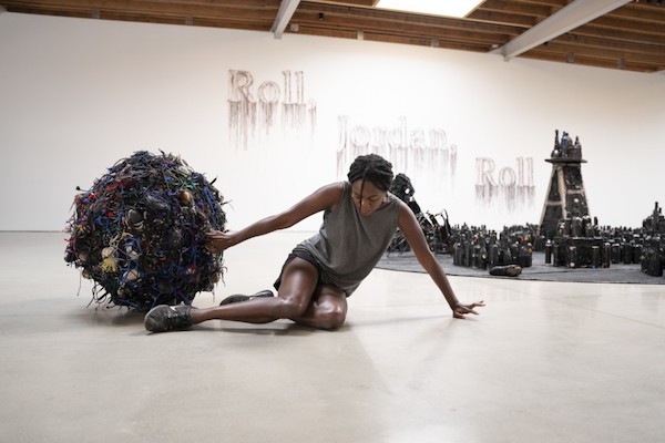 A woman sits on the ground in a white-walled room with the word "roll" on the wall in a descending diagonal. The woman has one hand on a large ball, an artwork called "Tumblehood" and behind her is more sculptural works made of sneakers and shoelaces. 