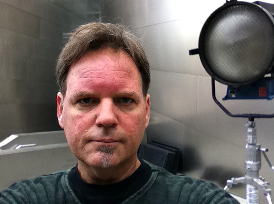 A man, Curtis Hayes, looks seriously into the camera, a big spotlight unlit behind him.