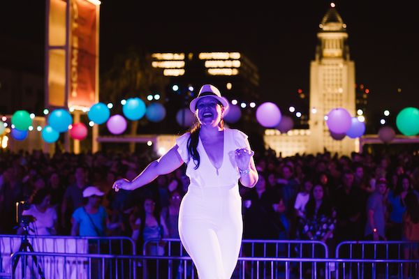 A woman dressed in white dances at night among colorful balloons with the LA city hall in the background
