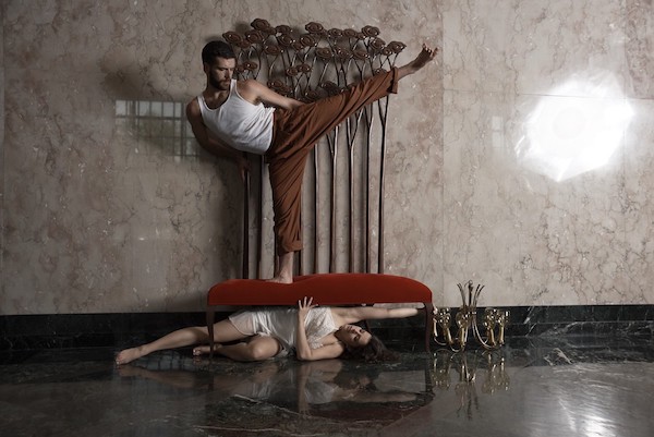 Photo of dancer standing on couch with another dancer stretched out beneath, her hand touching a chandelier on the floor.