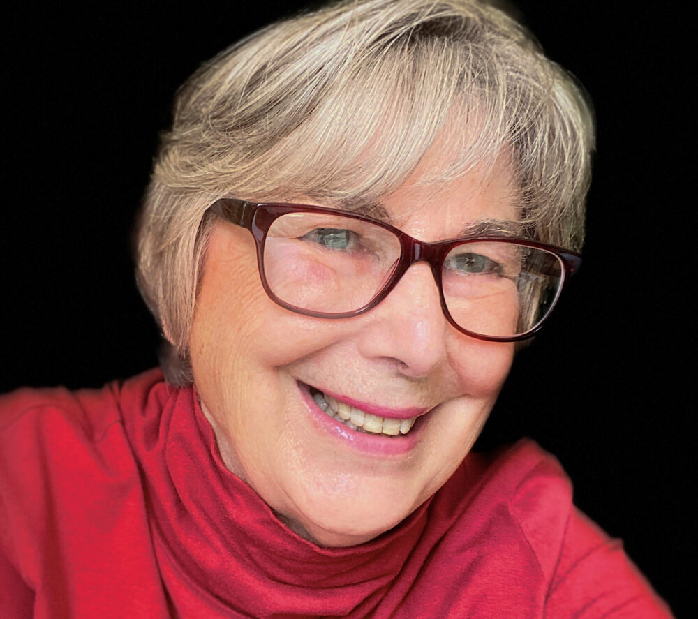 Headshot of Jackie Craven in red turtleneck shirt with a black background. She has glasses, short hair, in her 60s, smiling to the right of the camera.