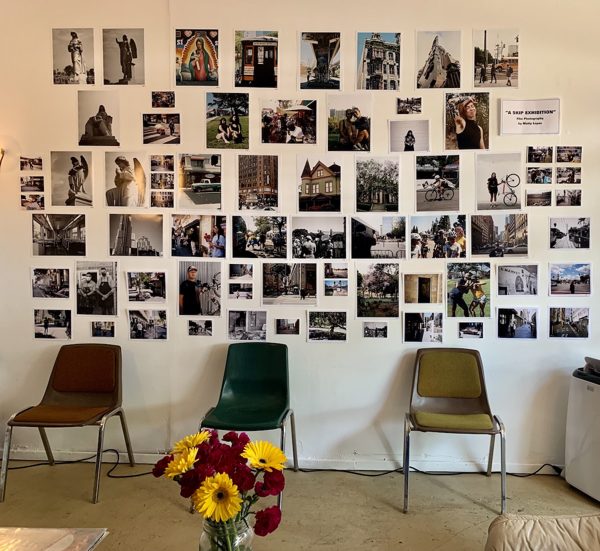 About 50 photos displayed frameless on a white wall, above three plastic chairs.