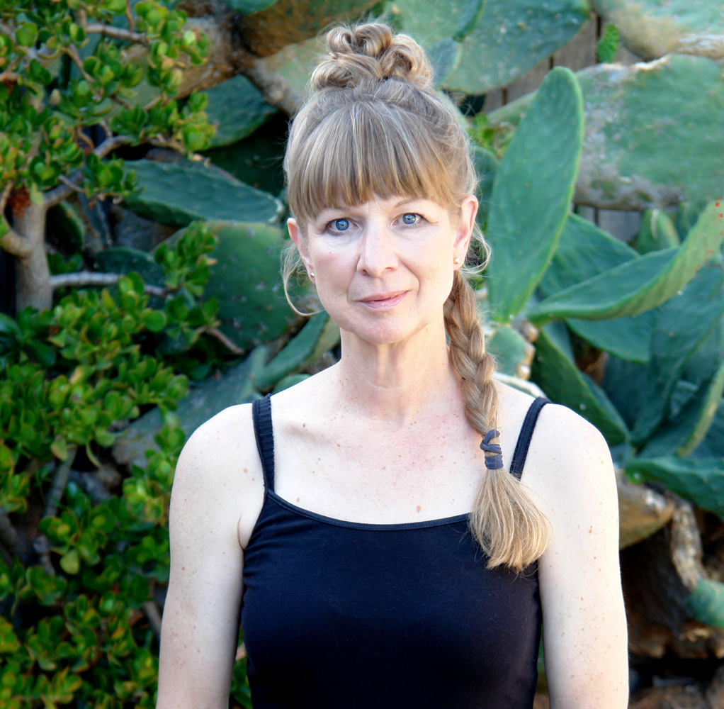 A woman, Wendy Rainey, her blond hair tied in a long braid, with a black strappy top, stands in front of large cacti.
