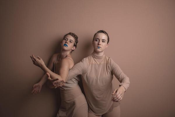 Two young female dancers in neutral clothing and blue lips look toward the camera.
