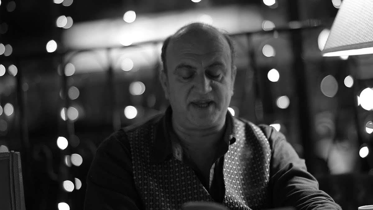 a black and white photo of a man reading from a book, lights sparkling behind him. this is david dephy, creator of poetry orchestra.