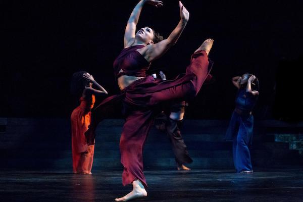 A dancer in red lifts her back leg toward her upstretched arms in front of other dancers