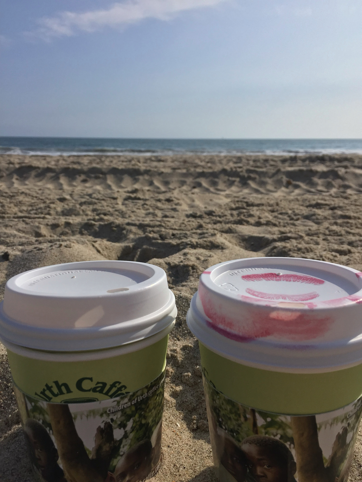Two to go coffee cups, one lid stained with lipstick, at the beach