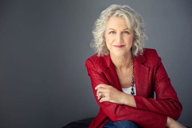 Photography of poet Nancy Murphy. She has on a red jacket, sitting confidently and staring directly at the camera.