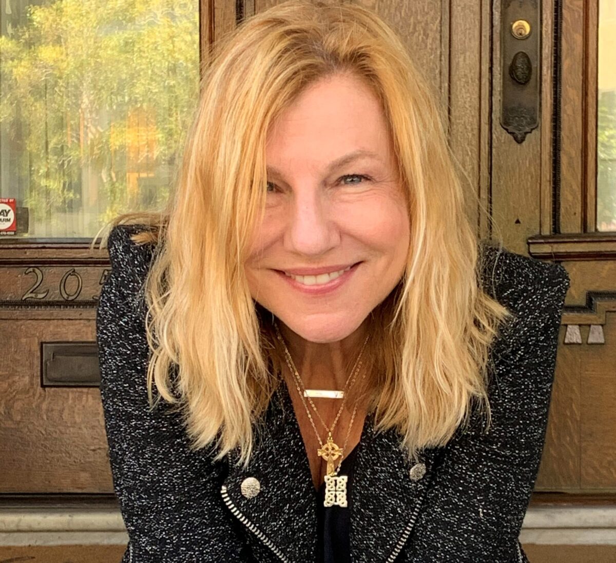 Photo of poet Valarie Hastings in jacket staring and smiling at the camera. Blonde haired, she has three pieces of jewelry dangling from her neck.