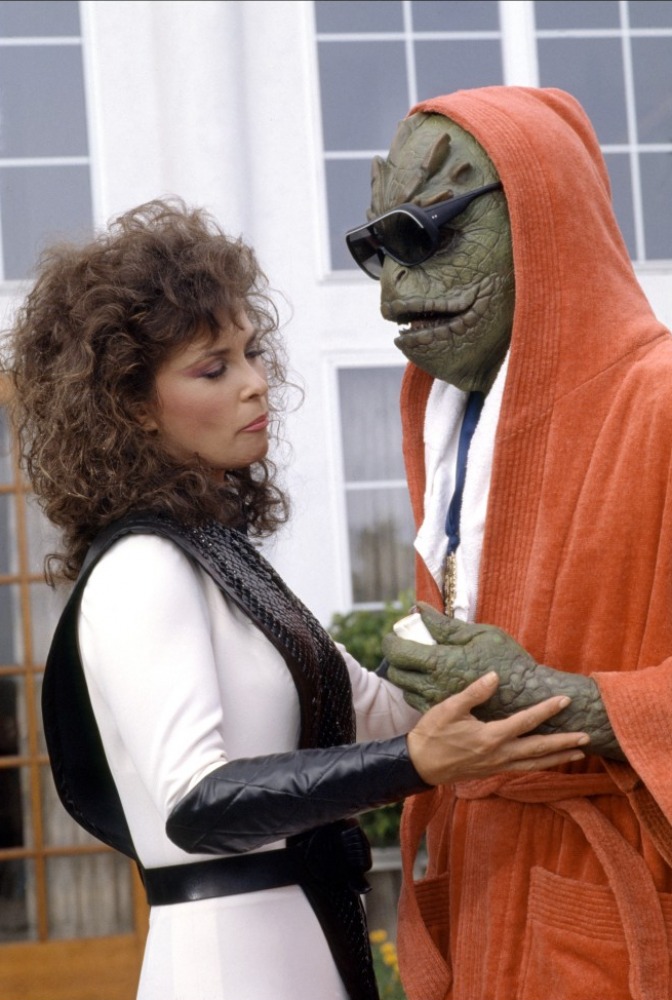 A lizard humanoid in an orange bathrobe and sunglasses with a woman in a white and black space jumpsuit.