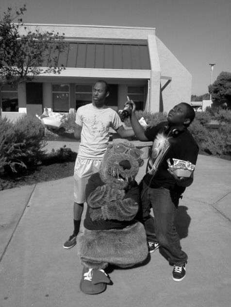 A black & white photo of two young Black men goofing around with a mascot.