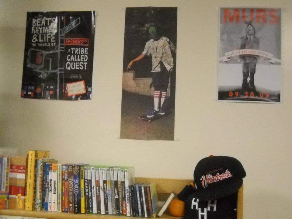 a dorm room, three posters on wall above a small bookcase that holds books and hats.