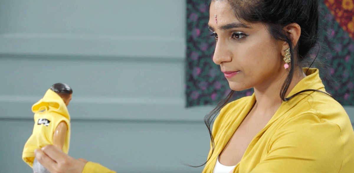 A moment in The Sitayana (Or How To Make An Exit), a woman in a yellow jacket staring at a doll in her hand, also dressed in a yellow outfit.