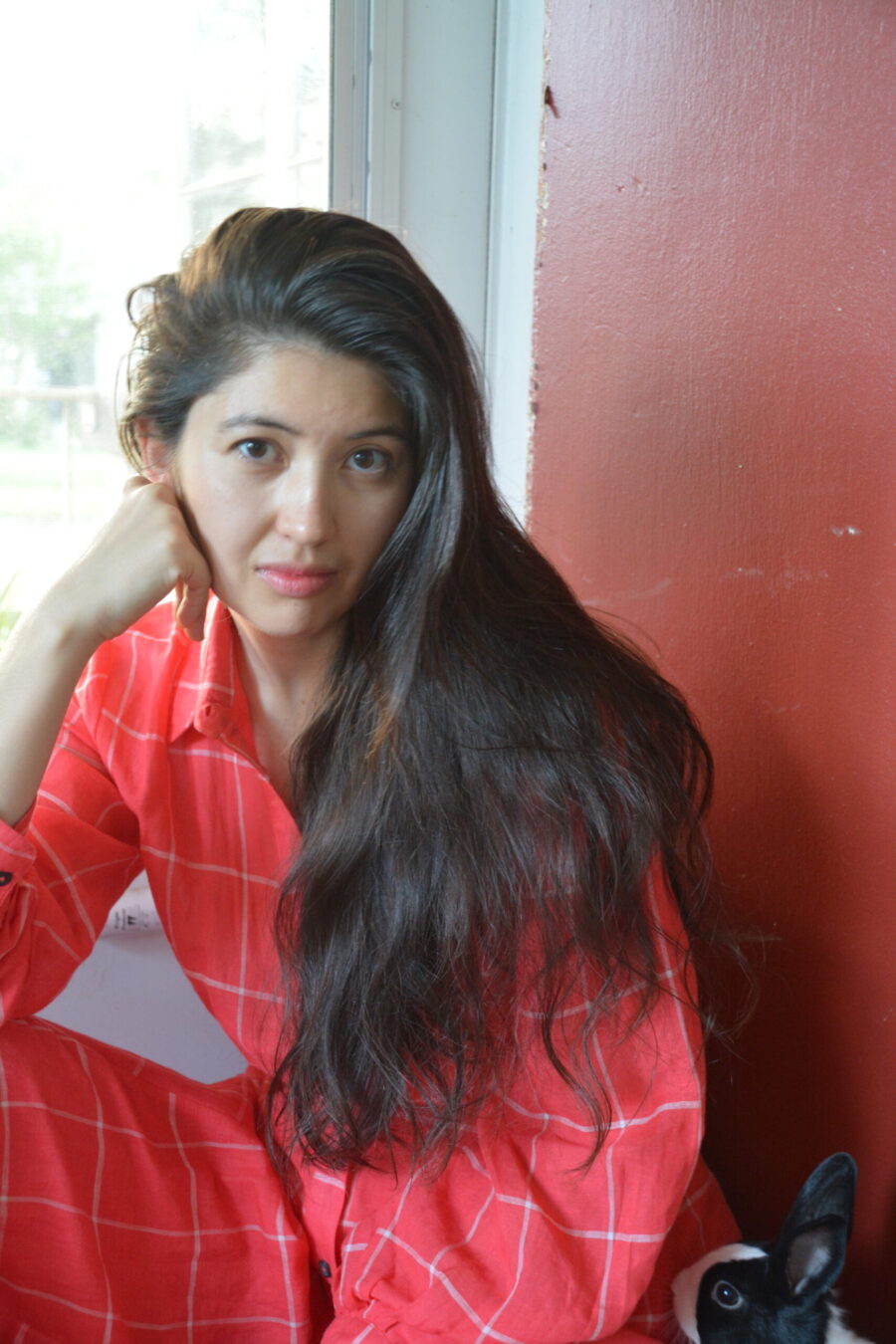 Color photo of Filipina poet Asa Drake. She's dressed all in red shirt and red pants, her long black hair hangs on her left shoulder. Her face resting on her hand, she stares directly at the camera.