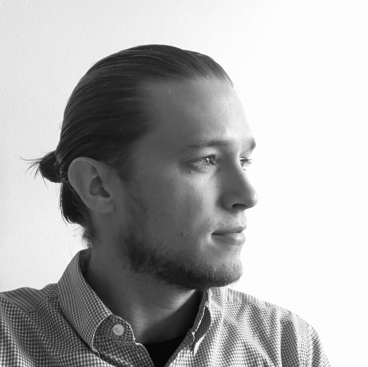 Black-and-white photo of poet Austin Velman starring to his left. He's in a checkered shirt and his hair is tied in a pony tail. He has a scruffy beard. He is trying to smile as he looks to his left away from the camera.