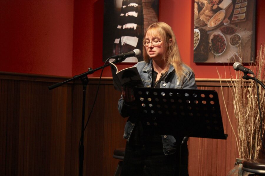 Photo of poet Rebecca Schumejda standing behind a poem. She's holding her book of poems and reading from it. She's on a stage and looks vulnerable.