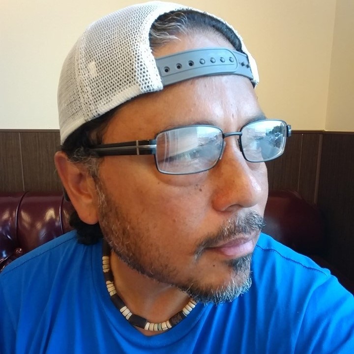 Photo of poet Tony Robles looking sideway. He wears a blue shirt, with glasses, and his hat on backward.