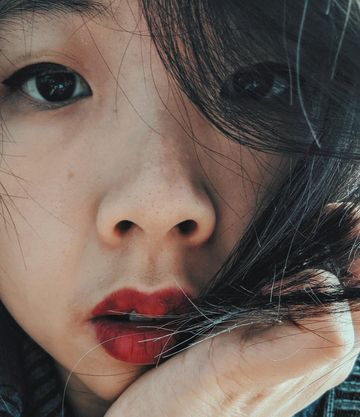 Cassandra Khaw, author of Nothing But Blackened Teeth, with red lipstick, her left hand holding a clump of her hair in front of her face