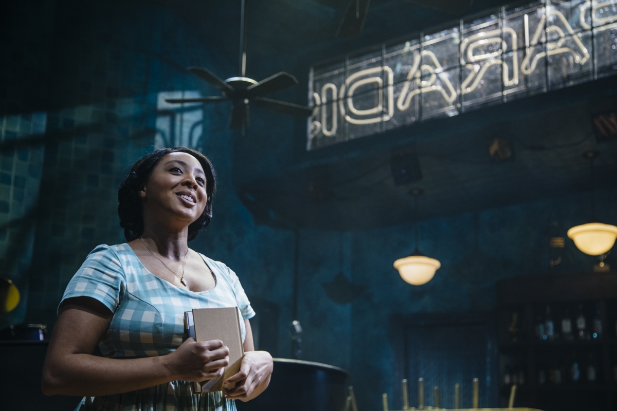 From a moment in Paradise Blue, a young Black woman holds a book, looking up and smiling, in a blue checkered dress in front of a sign.