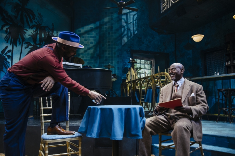 From a moment in Paradise Blue, two Black men, one sitting in a gray suit, the other in a red shirt and blue pants and hat, his left foot on a chair, pointing with a glass in his hand.