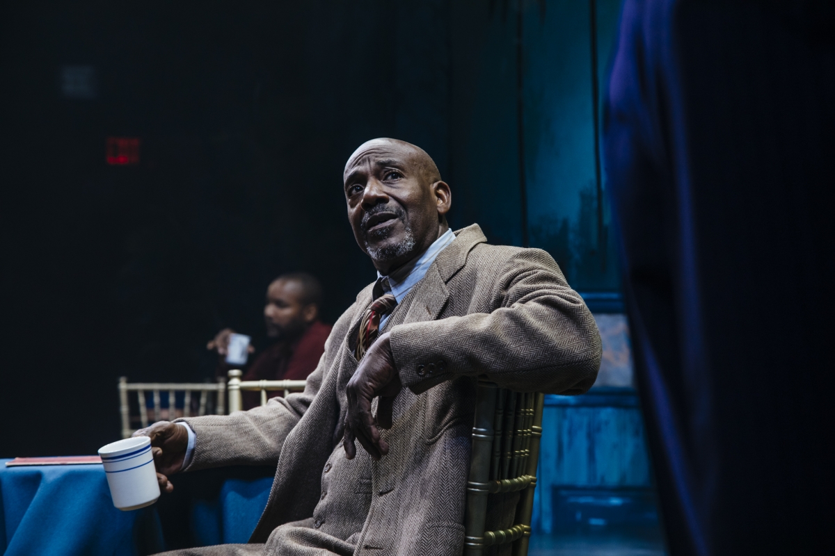 From a moment in Paradise Blue, a Black man in a gray suit, a coffee mug in his left hand, looks up with exasperation on his face.