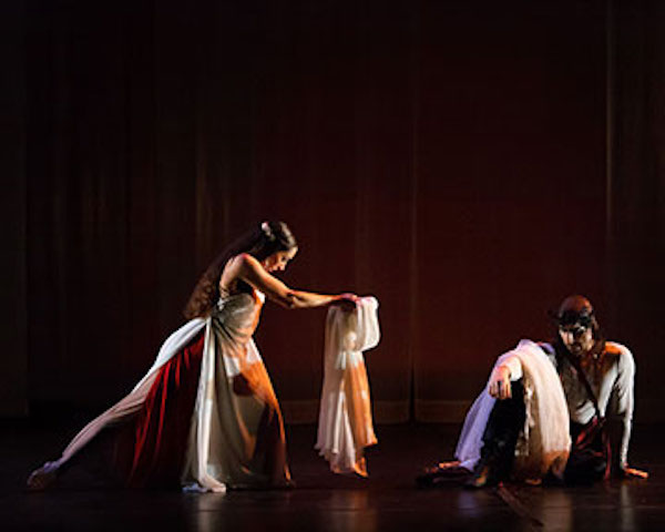 A woman bows and extends fabric to a sitting man