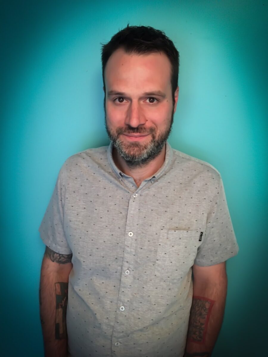 A photo of poet John Sibley Williams from the waist up. He has grey in his beard. Tattoos on both arms. Wears a grey shirt, looking directly at the camera and smiling.