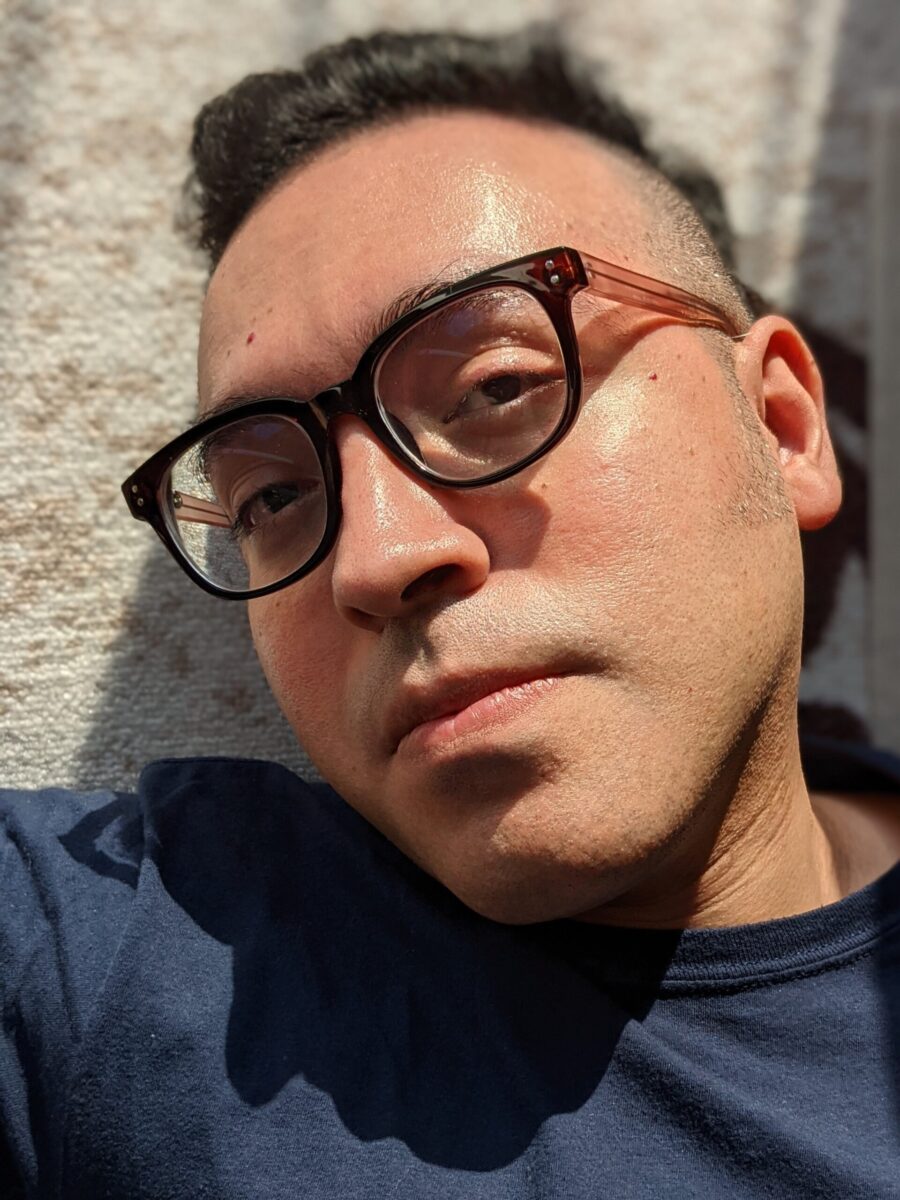 A headshot of poet Jose Araguz. He has on a black-rimmed glass, head titling, eyes looking at the camera, not necessarily smiling. It looks like the sides of his head are shaved.
