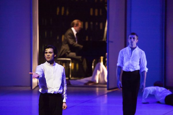 Two men stand in front of a man sitting at the back of the stage