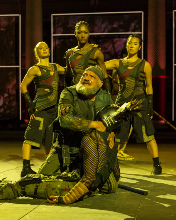 An old man sits in front of three warrior dancers