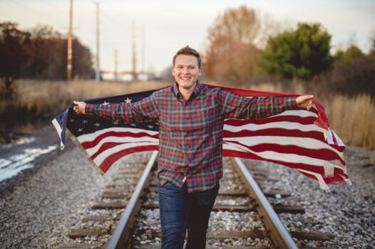 A smiling male holding the United States flag while walking on the train rails