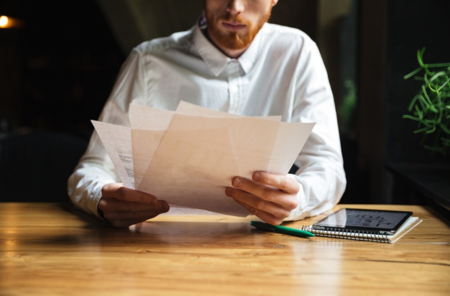 Man with red beard in white shirt reviewing papers at a wooden desk with a notebook and pen nearby